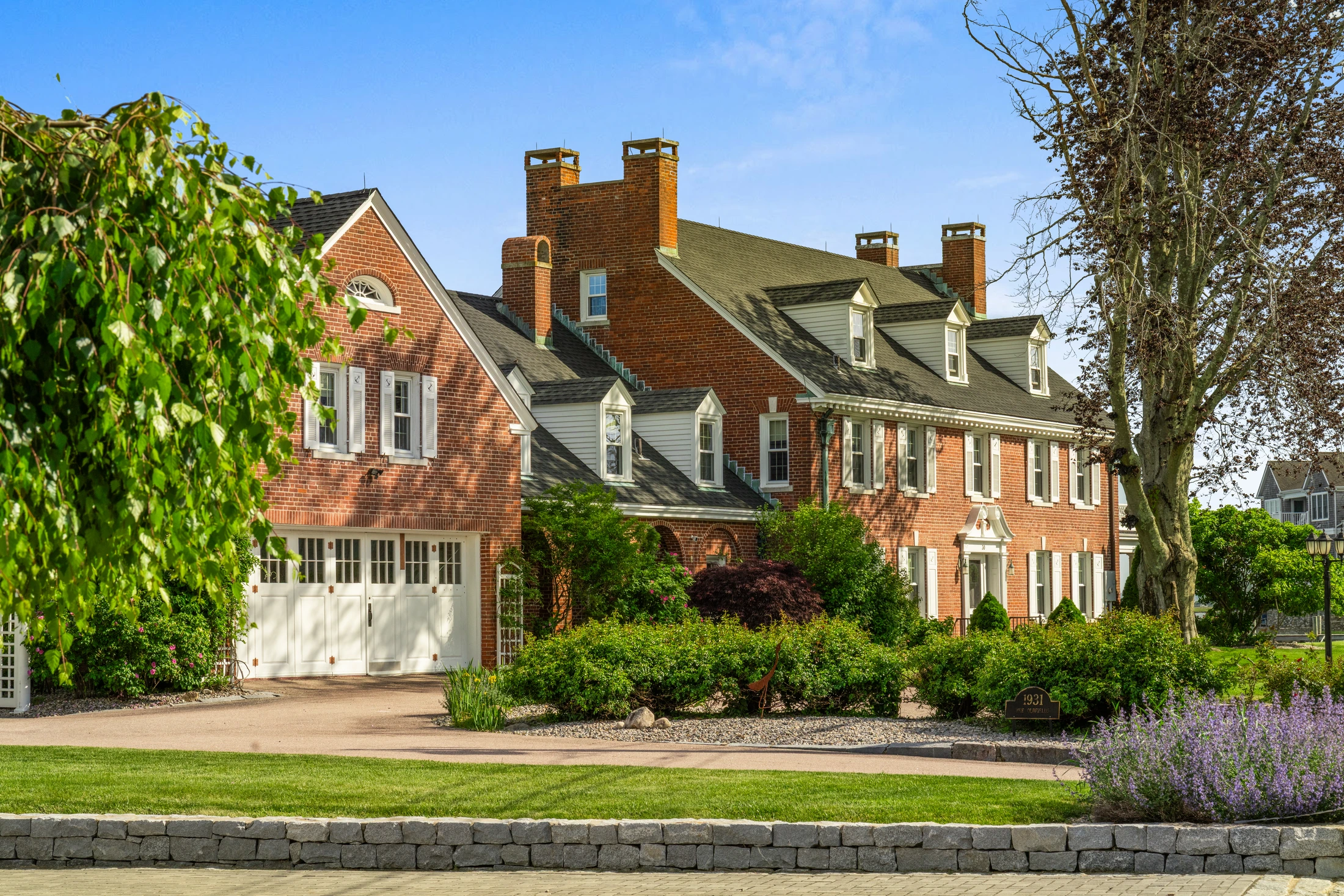 Drive approach to the brick residence with garage bay, gardens, and dormered roofline.